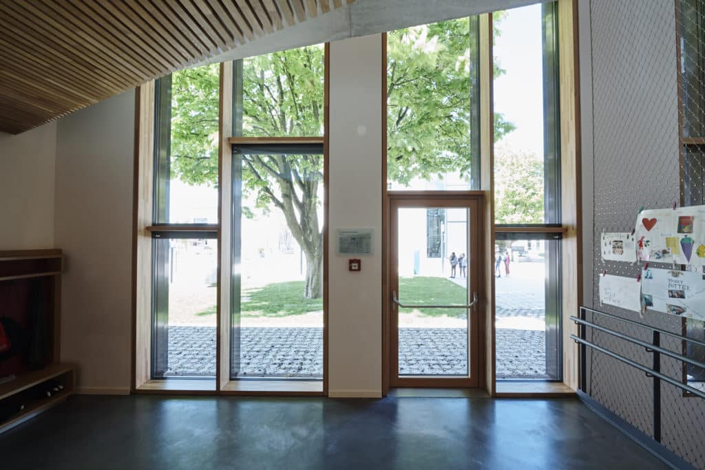 Eingangsbereich mit einer hohen Wand aus Fenster. Die Fensterrahmen sind aus massivem Holz