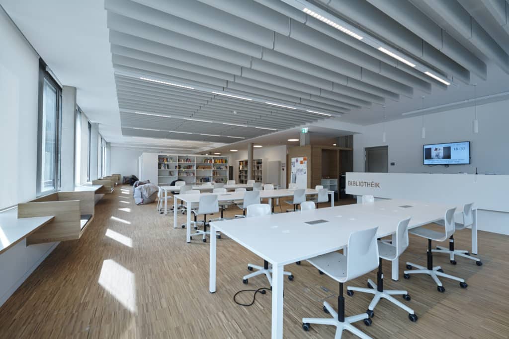 Klassenraum mit Boden aus Perkett. Sitzbänke aus Holz und Tische in weiß.