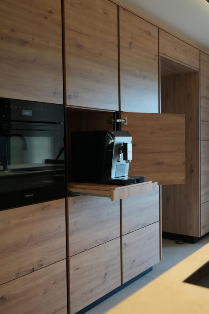 Photo d'un mur avec des armoires d'une cuisine à l'aspect bois. On voit le four et une armoire ouverte avec une tablette coulissante avec une machine à café.