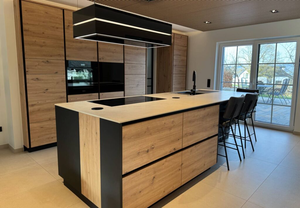 Gesamtaufnahme einer Küche in Holzoptik mit schwarzen Details. Freistehende Kücheninsel mit Waschbecken und Herd. Schwarze Abzugshaube.